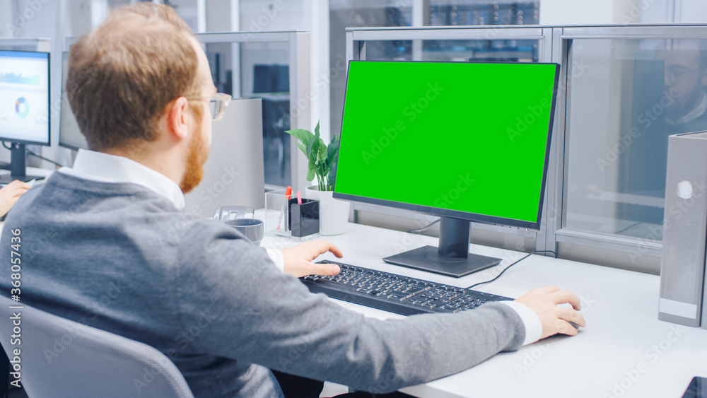 Poster Over the Shoulder Shot of Motivated Businessman Working on Green Mock-up Screen Desktop Computer in the Big Bright Open Space Office