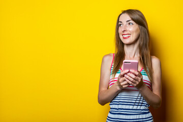 Young girl in a striped dress with a phone smiling looks to the side on a yellow background. Banner.