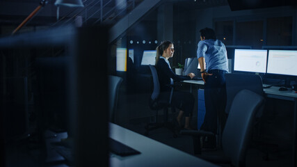 Confident Businessman Uses Desktop Computer, Her Male Manager Explains Specific Tasks, Account Handling and Strategy. Professional People Working Late at Night in Corporate Office