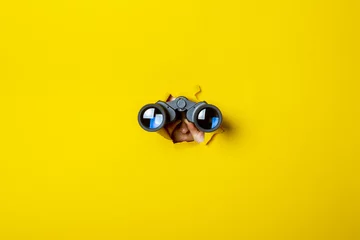 Fotobehang Female hand holds black binoculars on a yellow background. Journey, find and search concept © Alex