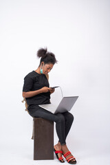 an african girl wearing a face mask, carrying a backpack, sitting and using her mobile phone and laptop