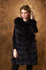 Young beautidul glamorous woman in black luxury expensive fur waist coat posing against golden wall. Fashion portrait