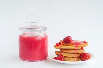 A jar of pink jam made of red currants and strawberries and a plate of pancakes.