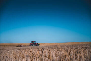Fototapeta premium Red tractor with plow on the field. Tractor on the field. Summer field and red tractor. Blue sky in background.