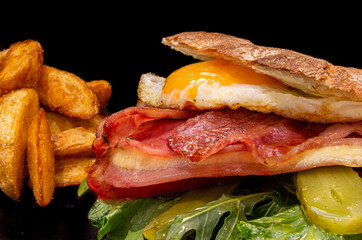 Primer plano de hamburguesa / sandwich con lechuga , tomate, huevo frito, bacon, jamon y queso, sobre fondo negro con patatas fritas.