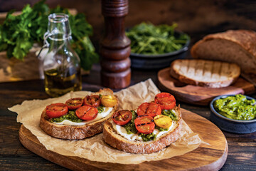 Bruschetta with cream cheese, pesto sauce and grilled cherry tomatoes, herbs on chopping board, rustic wooden table at home kitchen. Italian food, antipasto snack. Selective focus, copy space.