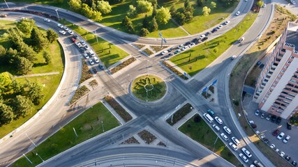 Aerial view of the junction road. There is a roundabout in the middle. Multi lane road, trees and buildings can be seen. Vehicles are waiting in the red light. There is summer and sunset light. 