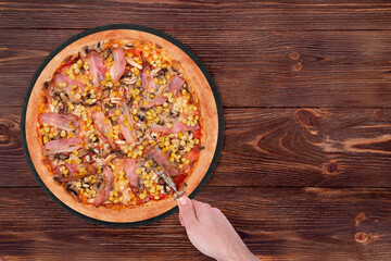 Man cuts the tasty pizza with chicken breast, corn, bacon and mushrooms, with a pizza cutter on a black round plate which is on wooden rustic table, copy space
