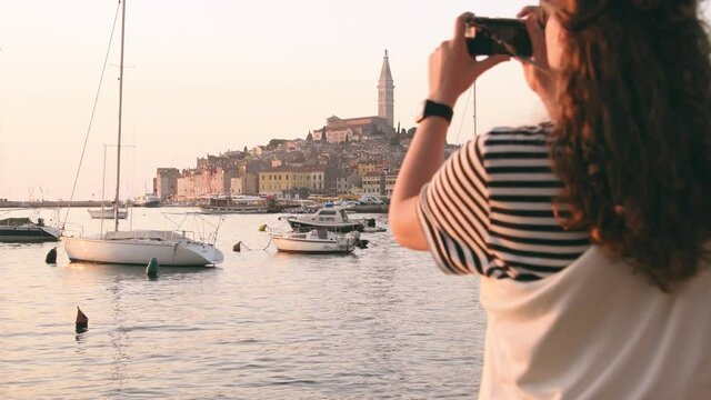 Young woman taking a picture of Rovinj marina and city view with boats and St. Euphemia cathedral at sunset. Istria, Croatia.