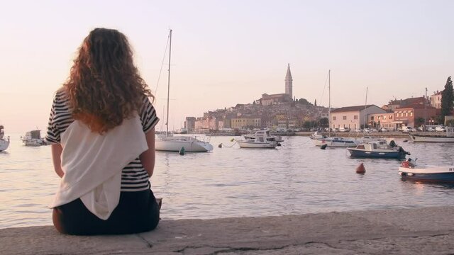 Young woman sitting on Rovinj marina and enjoying city view with boats and St. Euphemia cathedral at sunset. Istria, Croatia.