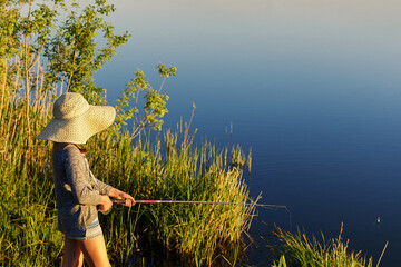 young girl in straw hat is fishing in the lake on summer evening from the shore overgrown with green tall grass