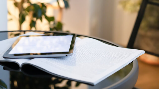 An open magazine and tablet on a glass black coffee table. Blurred bokeh background, close-up. Light interior with sunlight, sunset, bokeh.