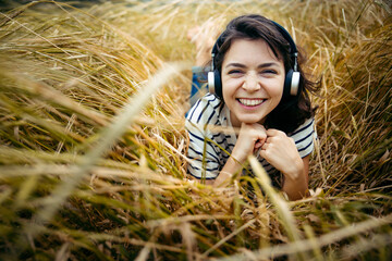 Cute hipster girl lies on the grass at wheat field and listening to music with iron headphones