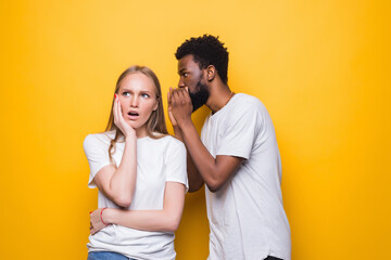 Cheerful young mixed couple whispering secret behind her hand sharing news posing isolated on yellow background. People lifestyle concept.