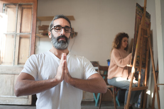 Man dressed in white sitting in a yoga pose with the hands together with a woman painting a picture in the background