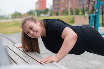 Beautiful fat young woman push up from the bench outdoors. A chubby smiling girl warms up before jogging. Sports for weight loss.