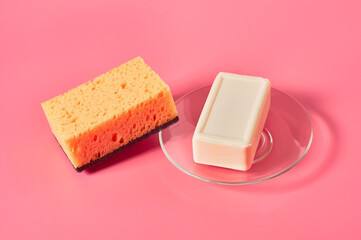 Brick of soap on glass saucer near sponge on pink background. Hygiene and cleaning concept