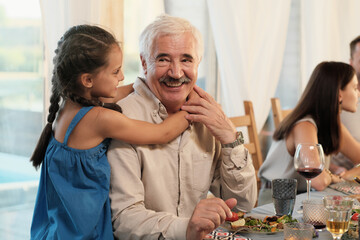Portrait of happy grandfather smiling at camera while his granddaughter embracing him during dinner at home