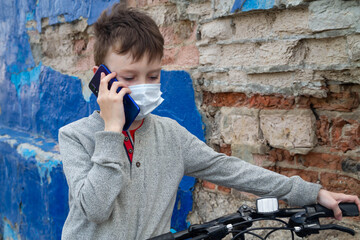 Obraz na płótnie Canvas A serious young European boy in a medical mask is talking on the phone with a friend and relative on the street near the blue wall.