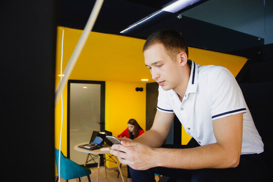 The guy is holding a smartphone. The office is in yellow and black shades. Social distance at work. The girl in the background works at the computer. - Powered by Adobe