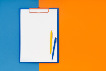 Blank clipboard mockup and two pens on blue-orange color background. Top view, copy space. Back to...