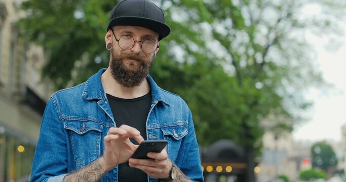 Portrait of handsome young Caucasian man in glasses and hat smiling at street. Stylish guy hipster with beard and ears tunnels chatting on smartphone and texting message tapping and scrolling on phone