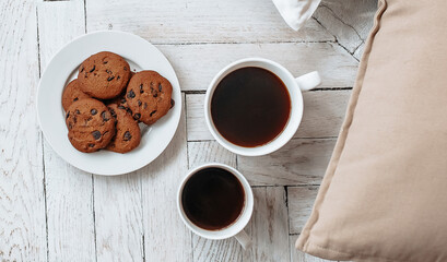 Breakfast in the morning. Two white cups of coffee and chocolate chip cookies stand on a light wooden parquet and pillows lie nearby. Homeliness, day off. Pastel colors.
