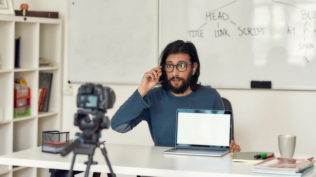 Young professional programming tutor recording video tutorial for his blog on professional digital equipment. Looking at camera and adjusting eyeglasses