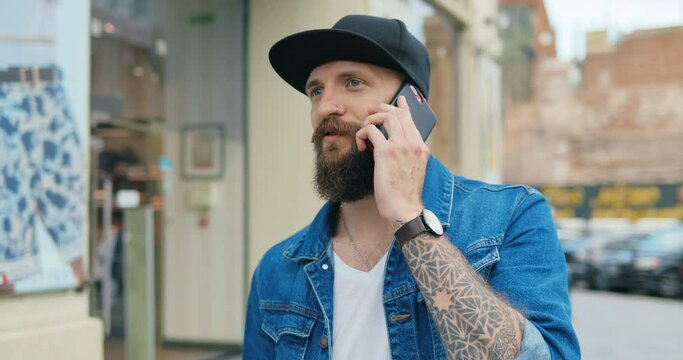 Caucasian handsome hipster man in hat and with beard strolling and talking outdoors on cellphone. Stylish guy speaking on mobile phone while walking at street.