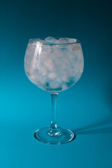 Head on view of a large gin glass of water and ice.  The glass is filled to the top with crushed and cubes of ice.  Set against a teal blue background with hard shadows.  Copy space available 
