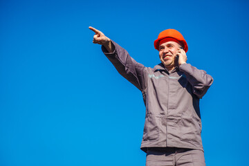 Construction worker in a helmet against the blue sky talking on the phone. Contractor control according to plan. Builder engineer helmet works at construction site. Labor day.