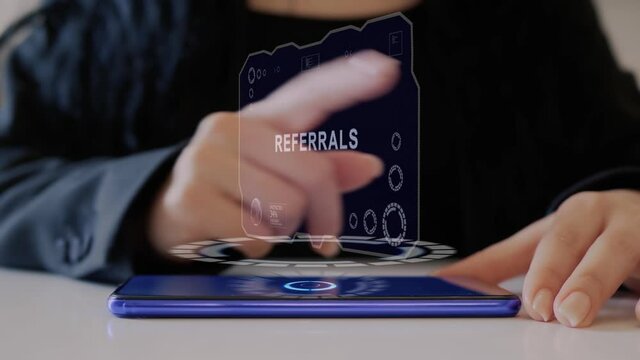 Female hand interacts HUD hologram with text Referrals. Woman in black uses conceptual holographic technology of the future on the screen of a smartphone lying on a table