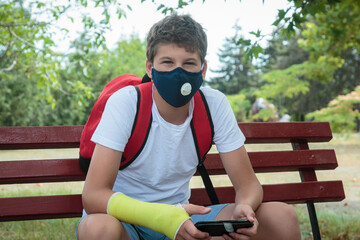 Close up young boy with protective mask on face on bench with phone. Teenager in white t shirt wearing medical mask to protect from corona virus, illness, infection. Quarantine on the city streets