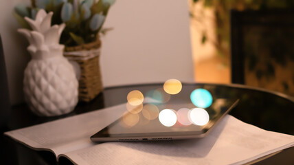 An open book and tablet on a glass black coffee table. Blurred bokeh background, close-up. Light interior with sunlight, sunset, bokeh.