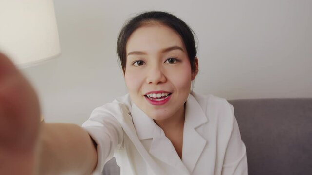 Cheerful beautiful Asian woman in a white shirt and look talk to camera at video calling recording with online chat on sofa at house.