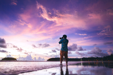 Rear view of adult travel asian photographer man with camera on beach sand with beautiful dramatic sunset sky - obrazy, fototapety, plakaty