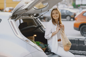 Charging electro car at the electric gas station. Woman sitting in a trunk. Lady with foodstuff.