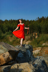 brunette girl with lush curly hair stands on the rocks near the lake in summer in a bright red dress with a violin, professional musician violinist