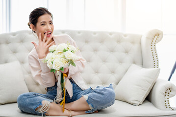 Beautiful charming Asian woman laying on white sofa and holding surprise white rose bouquet from her boyfriend at living room in the morning. Happiness relaxation mood concept.