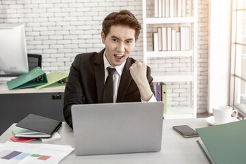 Fototapeta premium Happy of asian young businessman see a successful business plan on the laptop computer and pen on wooden table background in office,business expressed confidence embolden