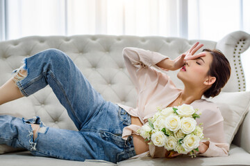 Beautiful charming Asian woman laying on white sofa and holding surprise white rose bouquet from her boyfriend at living room in the morning. Happiness relaxation mood concept.