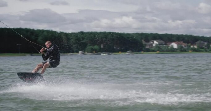 Wakeboarding in a lake near the forest, sportsman surfs on the water, ride on a wakeboarding board, performs tricks and does a somersault, 4k slow motion.