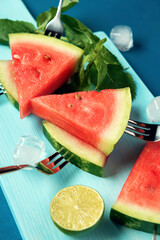 Watermelon slices with forks, mint, lime and ice cubes on the blue cutting board and the blue wooden background.
