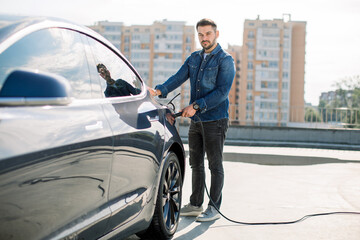 Full length portrait of young handsome bearded man in casual wear, standing at the charging station and holding a plug of the charger for an electric car. Eco electric car concept