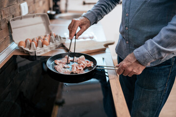 Senior man fries bacon in a pan at home.