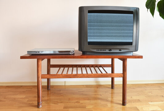 A DVD player and an outdated black TV with clutter on the screen sit on a vintage table in a 1990s apartment block.