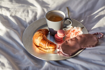 morning, hygge and breakfast concept - croissant, cup of coffee, macaroons and eye sleeping mask on plate in bed at home