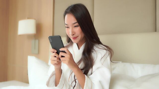 Portrait of Happy Asian Female Texting on Smarphone While Sitting on Bed in Bedroom