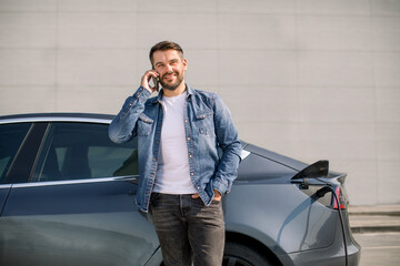 Smiling Caucasian man in casual wear talking phone while leaning on his luxury new electric car, standing on the charging station for charging a car. Electric car charging concept