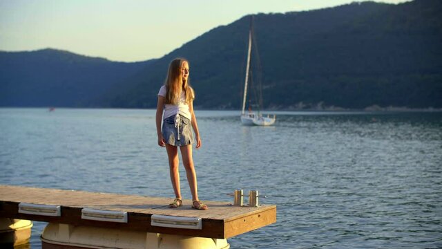 Tourist teen girl on sea pier looking around on green highlands and yacht background. Happy traveling girl admiring nature on lake pier on boat and mountain landscape